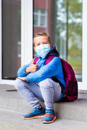 Cute looking at camera school boy 9s old sit on the stairs wears casual clothes and backpack face mask near school. Kid shows thumbs up image full lenghtの写真素材