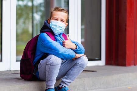 Cute looking at camera school boy 9s old sit on the stairs wears casual clothes and backpack face mask near school. Kid shows thumbs upの写真素材