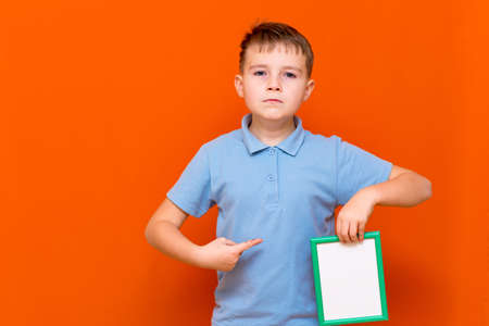 Portrait of caucasian schoolboy in t-shirt posing isolated on orange background. Mock up copy space. Shows to the white empty blank billboard with place for text or image.の写真素材