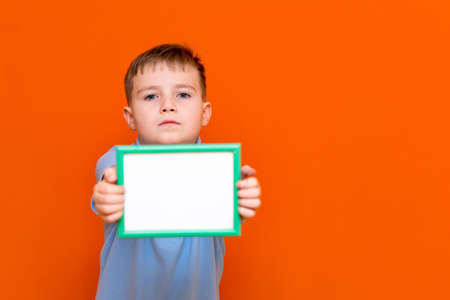 Portrait of caucasian schoolboy in t-shirt posing isolated on orange background. Mock up copy space. Shows advertising protruding forward white empty blank billboard with place for text or image.の写真素材