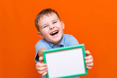 Portrait of caucasian schoolboy in t-shirt posing isolated on orange background. Mock up copy space. Shows advertising protruding forward white empty blank billboard with place for text or image.の写真素材