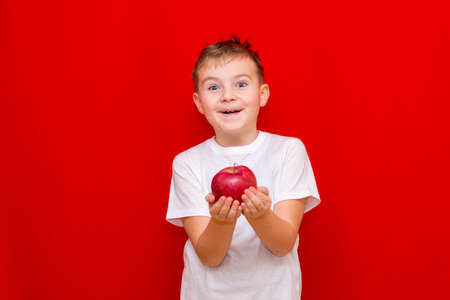 Close up European boy kid child schoolboy holds a red Apple oin his hands. Vitamins and fruits, healthy food. Prtrait of a happy beautiful child on a bright red background.の写真素材