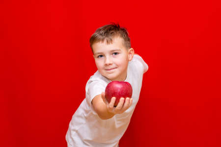 Close up European boy kid child schoolboy apple gives in front.Vitamins and fruits, healthy food. Prtrait of a happy beautiful child on a bright red background.の写真素材