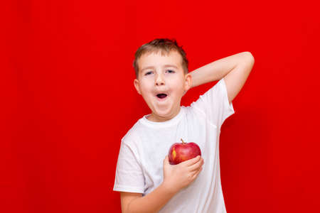 Close up European boy kid child schoolboy holds a red Apple oin his hands. Vitamins and fruits, healthy food. Prtrait of a happy beautiful child on a bright red background.の写真素材