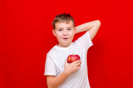 Close up European boy kid child schoolboy holds a red Apple oin his hands. Vitamins and fruits, healthy food. Prtrait of a happy beautiful child on a bright red background.の写真素材