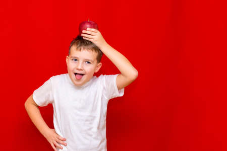 Close up European boy kid child schoolboy holds a red Apple on head .Vitamins and fruits, healthy food. Prtrait of a happy beautiful child on a bright red background.の写真素材