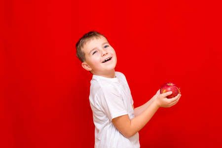 Side view Close up European smiling boy kid child schoolboy holds a red Apple oin his hands. Vitamins and fruits, healthy food. Prtrait of a happy beautiful child on a bright red background.の写真素材