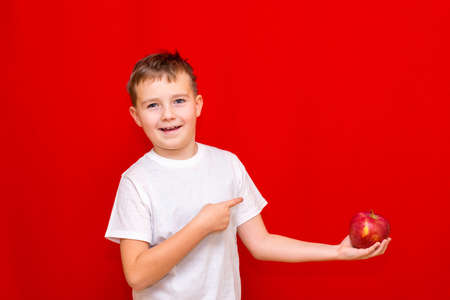 Side view Close up European smiling boy kid child schoolboy holds a red Apple oin his hands. Vitamins and fruits, healthy food. Prtrait of a happy beautiful child on a bright red background.の写真素材