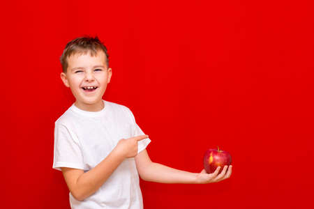 Side view Close up European smiling boy kid child schoolboy holds a red Apple oin his hands. Vitamins and fruits, healthy food. Prtrait of a happy beautiful child on a bright red background.の写真素材