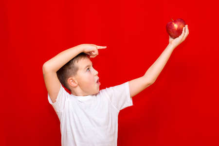 Close up European boy kid child schoolboy holds a red Apple oin his hands. Vitamins and fruits, healthy food. Prtrait of a happy beautiful child on a bright red background.の写真素材