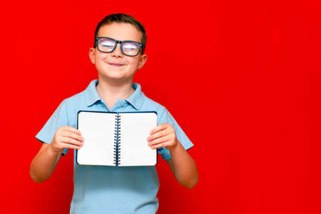 Cuccessful serious caucasian boy in a light blue t-shirt and glasses holds,shows with finger at blank notebook banner. Boy shows blank space. Bright red isolated studio wall.の写真素材