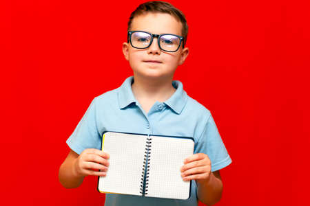 Cuccessful serious caucasian boy in a light blue t-shirt and glasses holds,shows with finger at blank notebook banner. Boy shows blank space. Bright red isolated studio wall.の写真素材