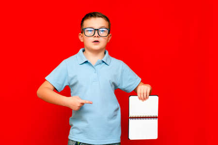 Cuccessful serious caucasian boy in a light blue t-shirt and glasses holds,shows with finger at blank notebook banner. Boy shows blank space. Bright red isolated studio wall.の写真素材