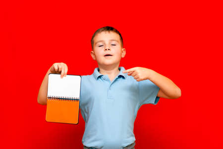 Strange cuccessful smiling calm, caucasian boy in a light blue t-shirt holds,shows with finger at blank notebook banner. Boy shows blank space. Bright red isolated studio wall.の写真素材