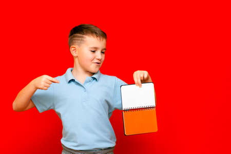 Strange cuccessful smiling calm, caucasian boy in a light blue t-shirt holds,shows with finger at blank notebook banner. Boy shows blank space. Bright red isolated studio wall.の写真素材