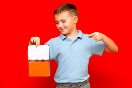 Strange cuccessful smiling calm, caucasian boy in a light blue t-shirt holds,shows with finger at blank notebook banner. Boy shows blank space. Bright red isolated studio wall.の写真素材