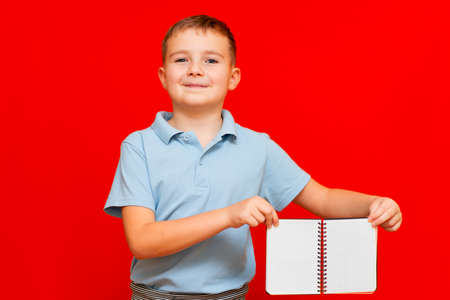 Strange cuccessful smiling calm, caucasian boy in a light blue t-shirt holds,shows blank notebook banner. Boy shows blank space. Bright red isolated studio wall.の写真素材