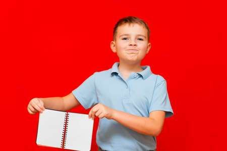 Strange cuccessful smiling calm, caucasian boy in a light blue t-shirt holds,shows blank notebook banner. Boy shows blank space. Bright red isolated studio wall.の写真素材