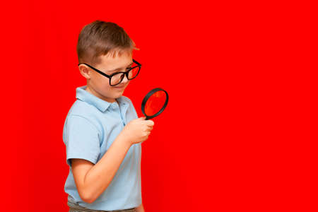 Side view Beautiful european young boy with eye glasses holding and looking through magnifying glass.Red studio wall.の写真素材