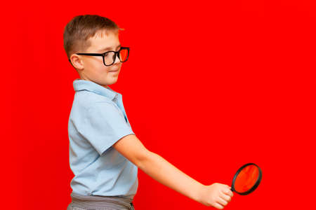 Side view Beautiful european young boy with eye glasses holding and looking through magnifying glass.Red studio wall.の写真素材