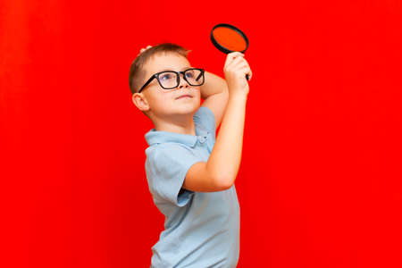 Beautiful european young boy with eye glasses holding and looking through magnifying glass somewhere .Red studio wall.の写真素材
