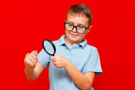Beautiful european young boy with eye glasses holding and looking through magnifying glass at his fingers palm fist .Red studio wall.の写真素材