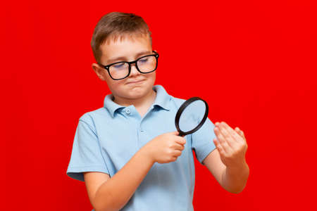 Beautiful european young boy with eye glasses holding and looking through magnifying glass at his fingers palm fist .Red studio wall.の写真素材