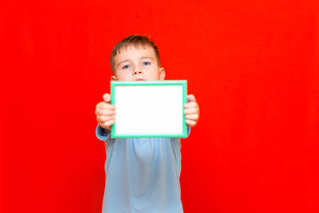 Happy small 9s boy holding in front a blank board advertisement mock up on red trendy bright background.の写真素材