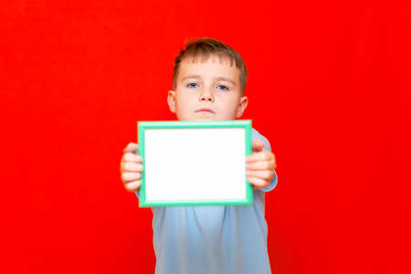 Happy small 9s boy holding in front a blank board advertisement mock up on red trendy bright background.の写真素材