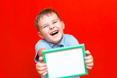 Happy small 9s boy holding a blank board advertisement mock up on red trendy bright background.の写真素材
