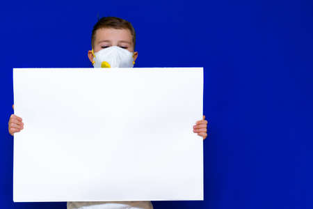 Close up Portrait of caucasian boy in a protective mask respirator holds a blank sheet of paper on a bright blue isolated background. Protection against coronavirus.Mock up,copy space,advertisement.の写真素材