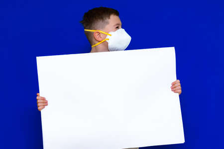 Side view Portrait of caucasian boy in a protective mask respirator holds a blank sheet of paper on a bright blue isolated background. Protection against coronavirus.Mock up,copy space,advertisement.の写真素材