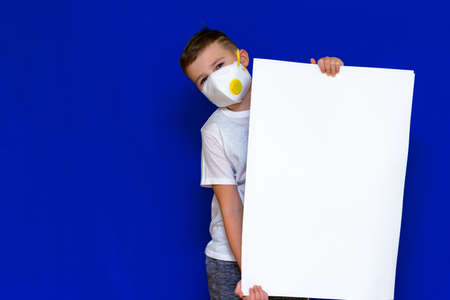 Close up Portrait of caucasian boy in a protective mask respirator holds a blank sheet of paper on a bright blue isolated background. Protection against coronavirus.Mock up,copy space,advertisement.の写真素材