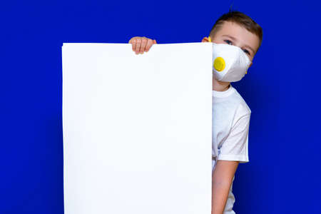 Close up Portrait of caucasian boy in a protective mask respirator holds a blank sheet of paper on a bright blue isolated background. Protection against coronavirus.Mock up,copy space,advertisement.の写真素材