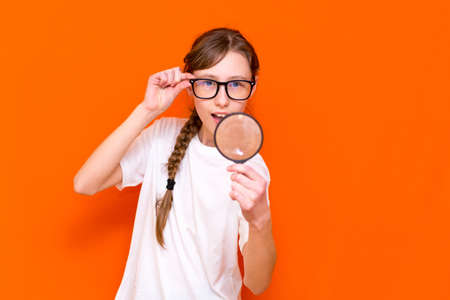 Attractive young girl in glasses looks through a magnifier on a bright orange background.の写真素材