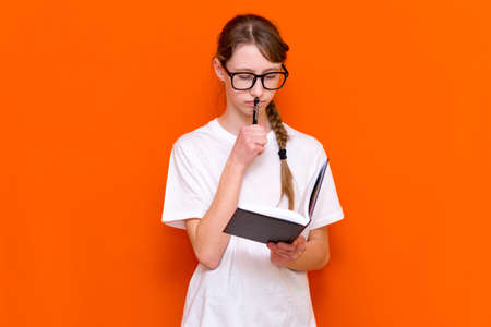 Excited thinking looking at camera young girl in eyeglasses, white T-shirt and pen something think and looking to a notebook on a orange studio background.Space for your text,banner,advertisement.の写真素材