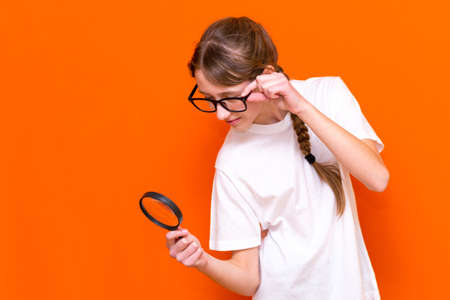 Attractive young girl in glasses looks through a magnifier on a bright orange background.の写真素材