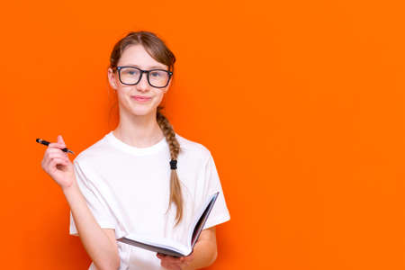 Excited thinking looking at camera young girl in eyeglasses, white T-shirt and pen something think and looking to a notebook on a orange studio background.Space for your text,banner,advertisement.の写真素材