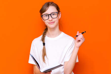 Excited thinking looking at camera young girl in eyeglasses, white T-shirt and pen something think and looking to a notebook on a orange studio background.Space for your text,banner,advertisement.の写真素材