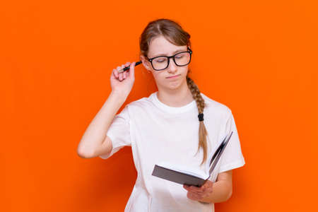 Excited thinking looking at camera young girl in eyeglasses, white T-shirt and pen something think and looking to a notebook on a orange studio background.Space for your text,banner,advertisement.の写真素材