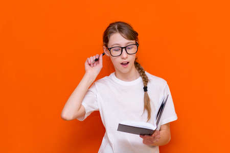 Excited thinking looking at camera young girl in eyeglasses, white T-shirt and pen something think and looking to a notebook on a orange studio background.Space for your text,banner,advertisement.の写真素材