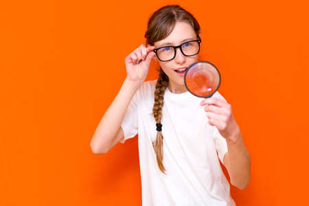 Attractive young girl in glasses looks through a magnifier on a bright orange background.の写真素材