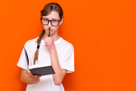 Excited thinking looking at camera young girl in eyeglasses, white T-shirt and pen something think and looking to a notebook on a orange studio background.Space for your text,banner,advertisement.の写真素材