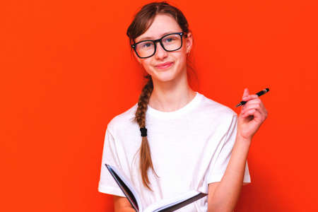 Excited thinking looking at camera young girl in eyeglasses, white T-shirt and pen something think and looking to a notebook on a orange studio background.Space for your text,banner,advertisement.の写真素材