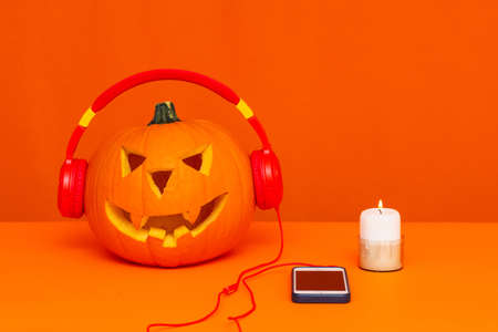 Carved pumpkin with headphones listening to music next to the phone and candle on the orange table bright orange backgroundの写真素材