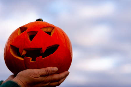 orange halloween carved pumpkin in woman hand with the blurred sky dark clouds background. Autumn...