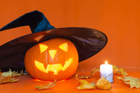 Jack o lantern with a black witch hat next to burning candle, on a orange table.Studio orange trendy...