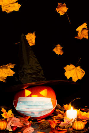Jack o lantern with a witch hat, on adark and yellow maple leaves background and with dry leaves, candle with medical face mask.の写真素材