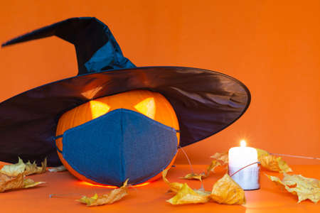 Jack o lantern with a black witch hat next to burning candle, on a orange table with protective mask.Studio orange trendy wallの写真素材