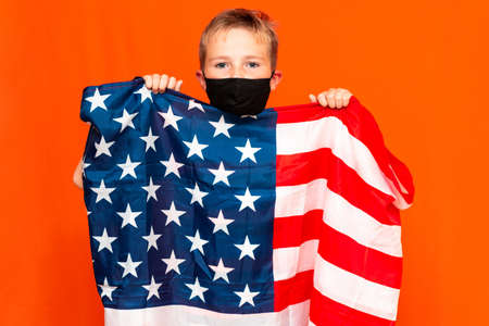 Young patriotic 9s child wears face mask holding united states of america flag on orange studio shot 4th of Julyの写真素材